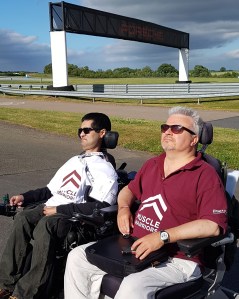[2 men in wheelchairs wearing Muscle Warrior t-shirts looking across to Silverstone race-track]